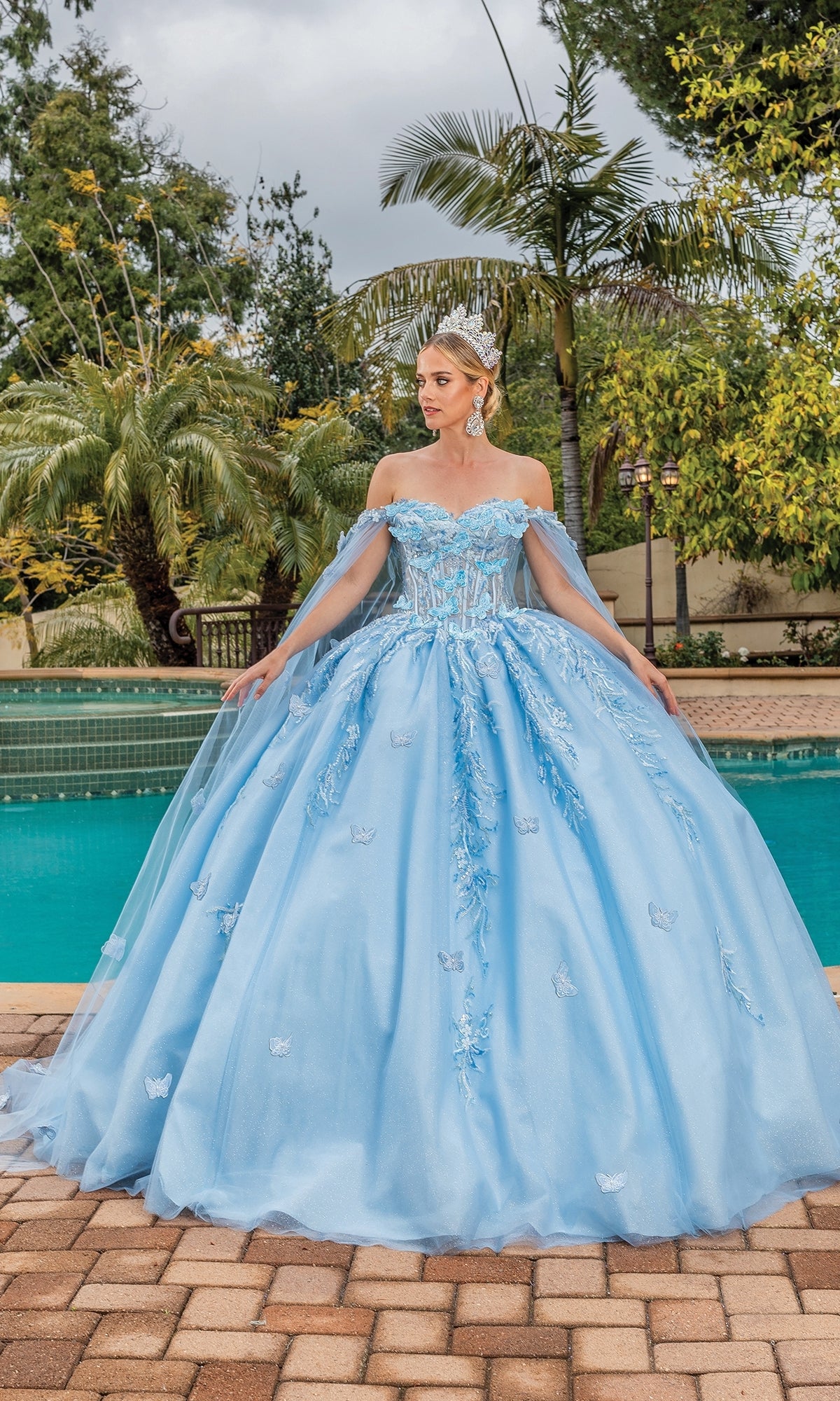 A woman in the Quinceanera Dress 1889 by Dancing Queen, a light blue off-the-shoulder ball gown, stands on a brick patio by a pool with palm trees and lush greenery in the background.